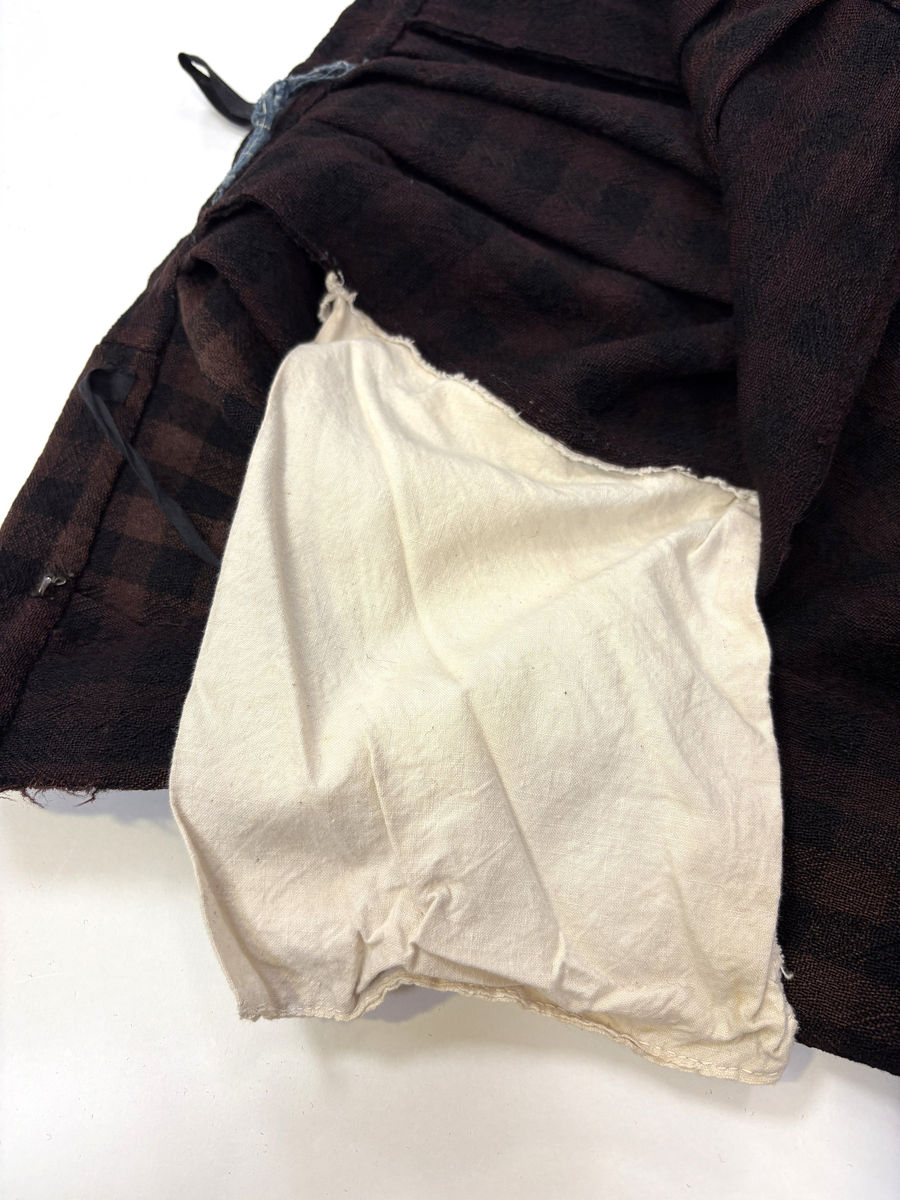 Stakk i samme stoff som liv SM.008023.01; svart og brunrutet ull i hjemmevevd toskaft. Stakken er falmet og slitt med en del hull spredt utover. Sydd sammen for hånd med attersting med svart bomullstråd av fem vevbredder som har jarekanter. Stakken har et 22 cm bredt glatt stykke midt på foran og foldelagt på begge sider. Åpningen er i venstre side og er 21 cm lang. Stakken har fire folder foldelagt mot høyre og seks folder foldelagt mot venstre, 3,5-4,5 cm brede. Midt på bak er det et 13 cm rynket felt med en halv fold over på begge sider. Foldene er tråklet med mørk blå ulltråd, sydd på med attersting med svart bomullstråd og så kastet med faldesting med svart bomullstråd. Linningen er i samme stoff som kjolen og sydd med faldesting med svart bomullstråd deretter med prikkesting med brun lintråd midt på linningen. Lukkes med to metallhekter. Stakken har en 4 cm bred hengefald 11,5 cm fra nedkanten, sydd med faldesting med svart bomullstråd, og en 7,5 cm bred skoning i stripet bomullsstoff sydd med faldesting med svart bomullstråd. Dette er kjøpetøy i fargene beige, rosa, lys blå, blå, mørk blå, hvit og grå. Stakken har en 21 cm lang og 19 cm bred lomme på høyre side i samme stoff som fôret på livet, sydd fast med attersting med ubleket bomullstråd. Stakken har en metallhempe midt på bak på linningen for å hekte på livet og skal brukes utenpå livet.