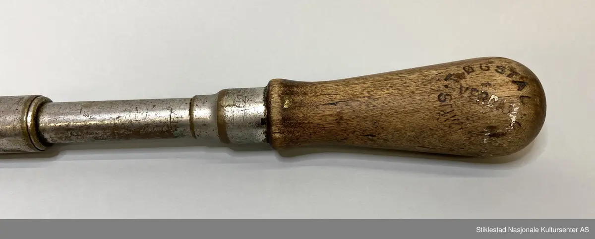 Skrujern med skrallefunksjon, flatskrujern. Skaft i løvtre, resten i metall/blikk. Utskiftbart skrujern, pumpefunksjon. Innskrift/stempel vanskelig å lese, men amerikansk. Skaft svimerket med T. Øgstad, Stiklestad, Verdal (rundt stempel)