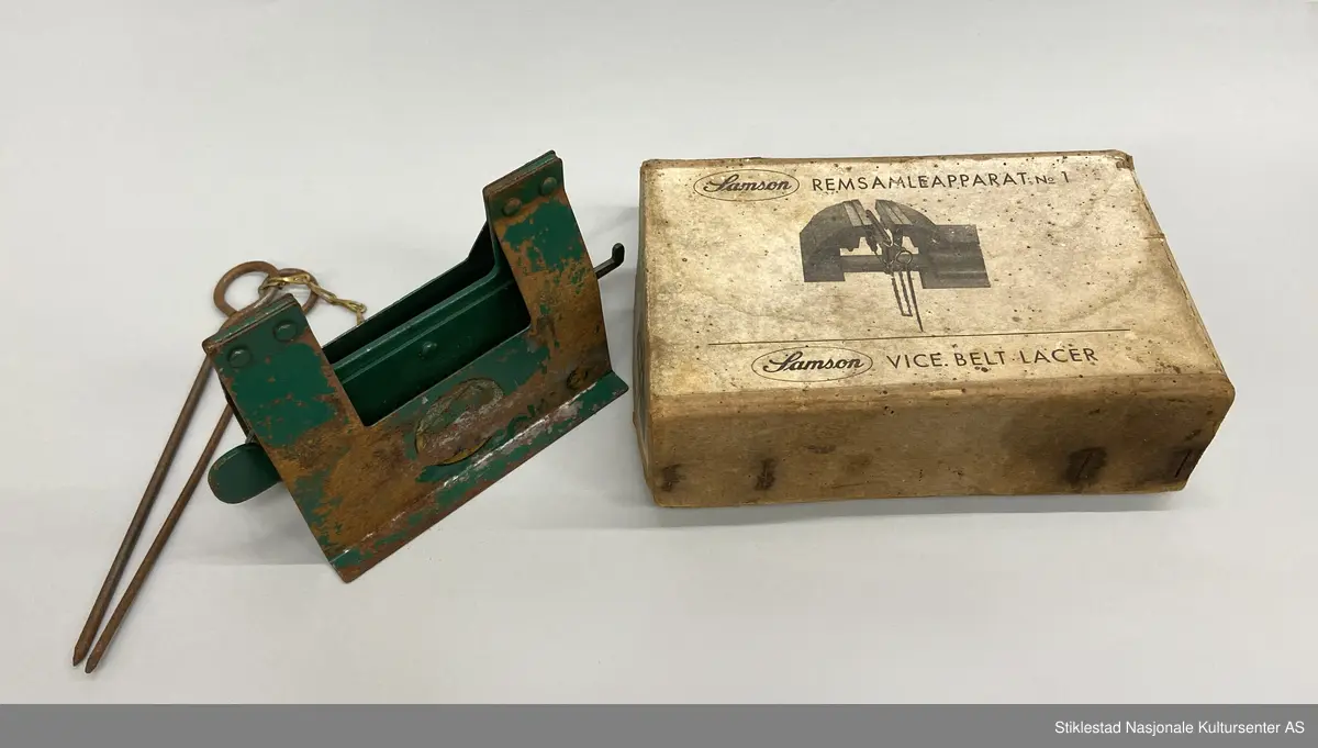 Eske med Samson Reimsamleapparat no 1/Samson Vice belt Lacer. Reimsamleapparat til skrustikke. Apparat i metall, grønnlakkert med prod.merke, uleselig. Mye rust. Fra apparatet henger det to «splinter»  fra en kort kjetting i messing. Esken er i kraftig papp og stiftet sammen. 3 deler. Skrevet med blyant på eska: 25,60 (pris?)
