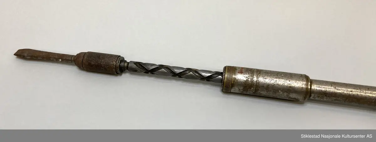 Skrujern med skrallefunksjon, flatskrujern. Skaft i løvtre, resten i metall/blikk. Utskiftbart skrujern, pumpefunksjon. Innskrift/stempel vanskelig å lese, men amerikansk. Skaft svimerket med T. Øgstad, Stiklestad, Verdal (rundt stempel)