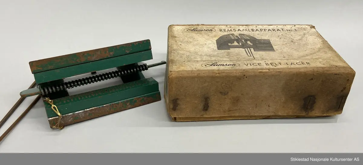 Eske med Samson Reimsamleapparat no 1/Samson Vice belt Lacer. Reimsamleapparat til skrustikke. Apparat i metall, grønnlakkert med prod.merke, uleselig. Mye rust. Fra apparatet henger det to «splinter»  fra en kort kjetting i messing. Esken er i kraftig papp og stiftet sammen. 3 deler. Skrevet med blyant på eska: 25,60 (pris?)