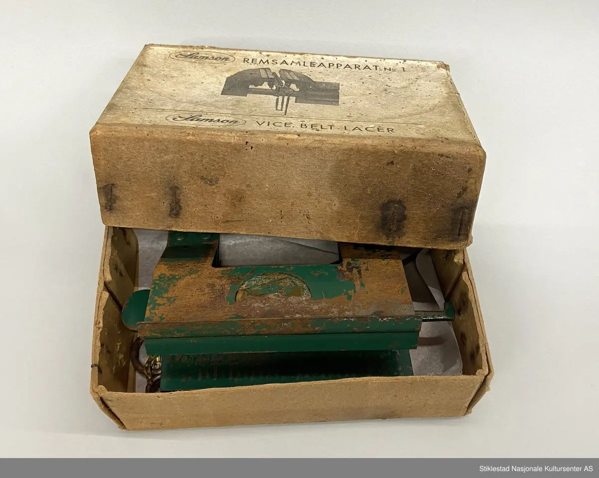 Eske med Samson Reimsamleapparat no 1/Samson Vice belt Lacer. Reimsamleapparat til skrustikke. Apparat i metall, grønnlakkert med prod.merke, uleselig. Mye rust. Fra apparatet henger det to «splinter»  fra en kort kjetting i messing. Esken er i kraftig papp og stiftet sammen. 3 deler. Skrevet med blyant på eska: 25,60 (pris?)