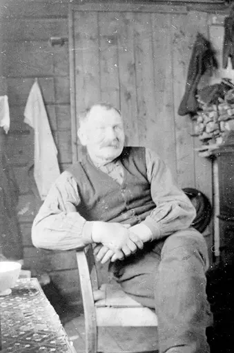 Portrett av en mann med bart som sitter på en stol ved et bord. Bak ham er det en dør, og på veggen henger det et håndkle.