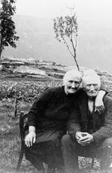 Mann og kone sitter i stol utendørs. I bakgrunnen fjell, træ