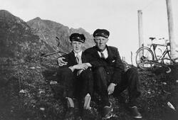 To menn sitter på bakken ved en innhegning. Tre sykler kan s