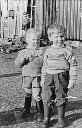 To gutter i høye støvler, ei jente ved husveggen i bakgrunne