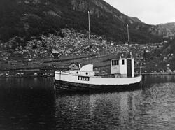 En fiskebåt på vannet, N 89 R. I bakgrunnen på land ser vi e