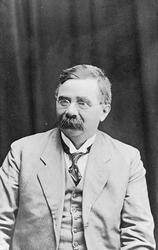 Portrett av en mann med briller og bart. Han har dress.