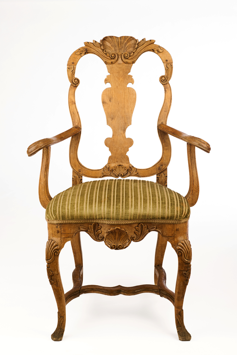 Stol av ask som hviler på fire ben. Gjennombrutt rygg bestående av C-former, samt symmetrisk svunget ryggbrett. På øvre og nedre del av ryggen symmetriske bladmotiv og skjell. Trapesformet sete, trukket med sekundært grønnfarget tekstil. Svungne armlener. Sargen er buet, innsvinget på midten og med utskåret skjellmotiv. Cabrioleformede forben med utskåret skjellornamentikk og klo og kule, samt svungne bakben. H-kryss mellom benene.