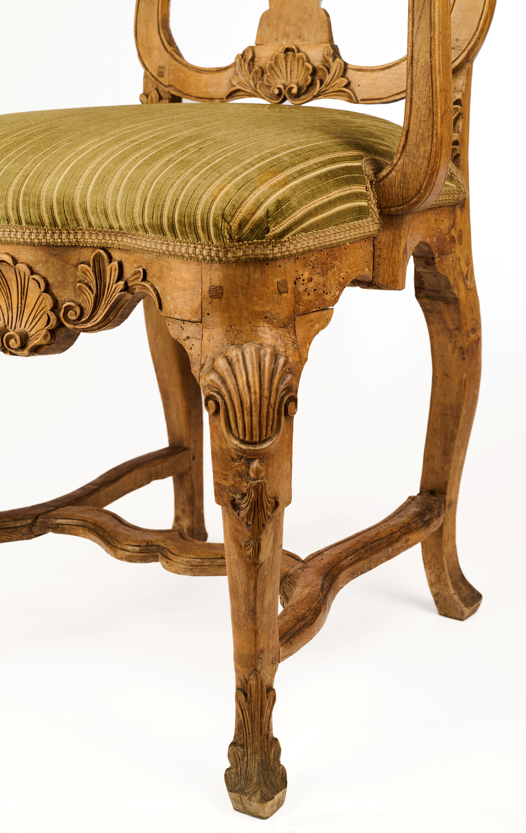 Stol av ask som hviler på fire ben. Gjennombrutt rygg bestående av C-former, samt symmetrisk svunget ryggbrett. På øvre og nedre del av ryggen symmetriske bladmotiv og skjell. Trapesformet sete, trukket med sekundært grønnfarget tekstil. Svungne armlener. Sargen er buet, innsvinget på midten og med utskåret skjellmotiv. Cabrioleformede forben med utskåret skjellornamentikk og klo og kule, samt svungne bakben. H-kryss mellom benene.