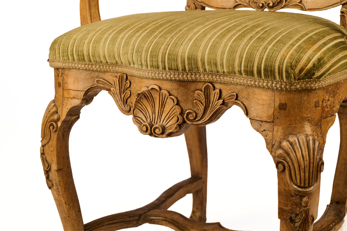 Stol av ask som hviler på fire ben. Gjennombrutt rygg bestående av C-former, samt symmetrisk svunget ryggbrett. På øvre og nedre del av ryggen symmetriske bladmotiv og skjell. Trapesformet sete, trukket med sekundært grønnfarget tekstil. Svungne armlener. Sargen er buet, innsvinget på midten og med utskåret skjellmotiv. Cabrioleformede forben med utskåret skjellornamentikk og klo og kule, samt svungne bakben. H-kryss mellom benene.