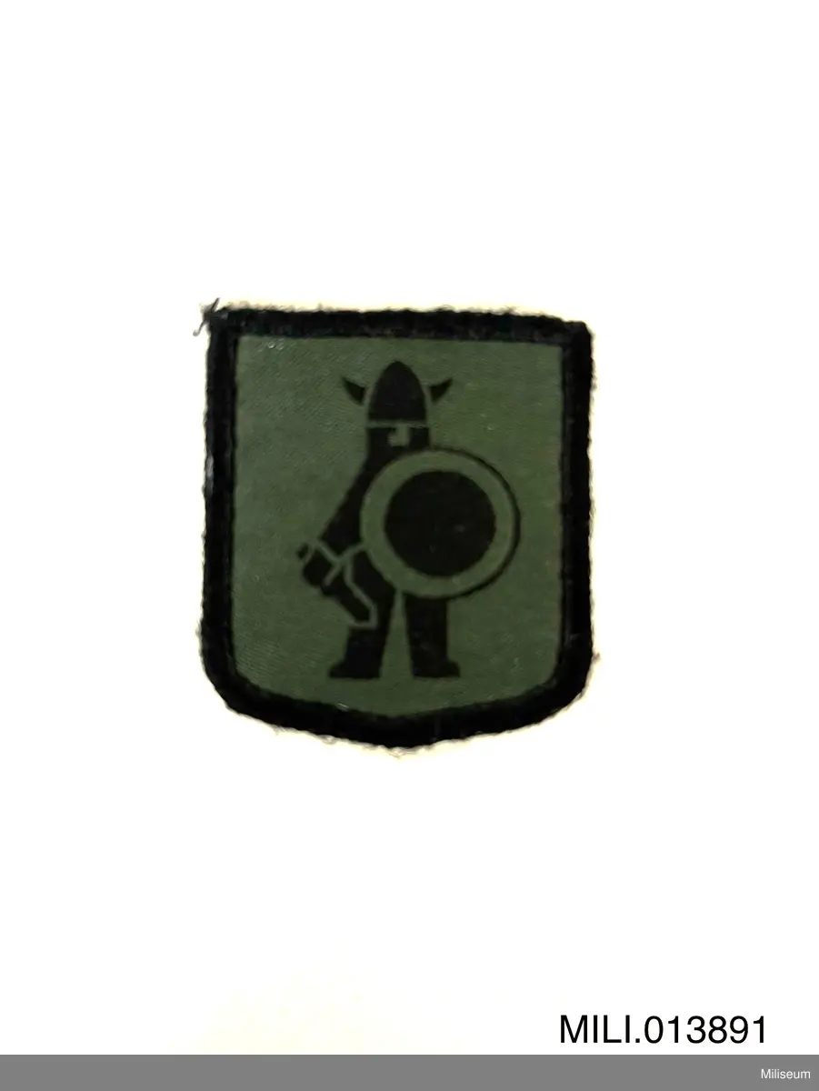 Tilläggstecken av textil med maskinvävt motiv. Med kardborrefästning på baksidan. Avsett att sys fästas på vapenrockens (motsv.) högra överärm.