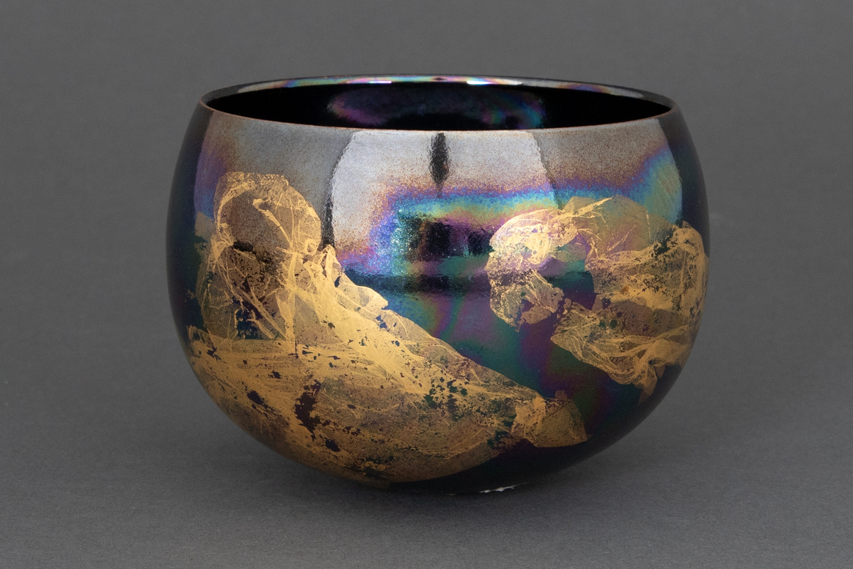 Bolle med liten bunnflate i porselen. Avrundet bunn og topp. Lilla, mørkeblå og grønn bunnkulør i bølgende formasjoner med uregelmessige former i bladgull utvendig.