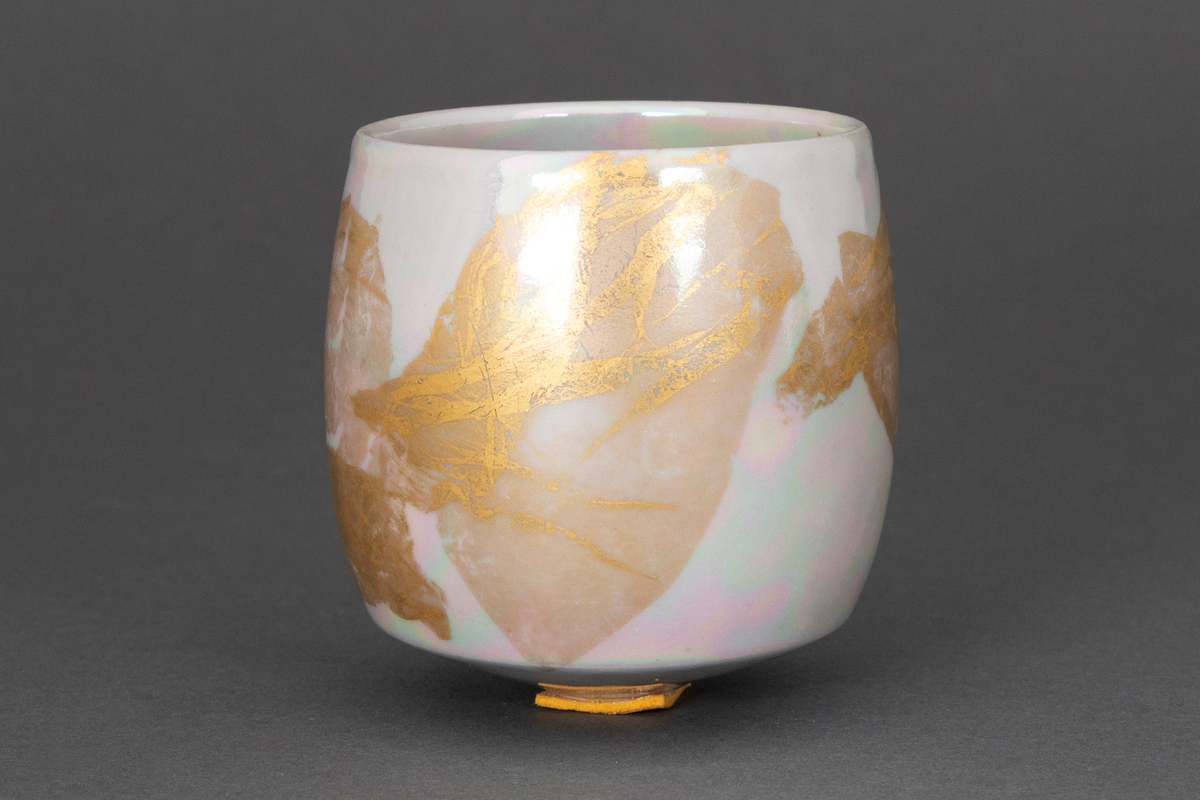 Bolle med liten bunnflate i porselen. Sylinderform som rundes av mot bunnen og toppen. Gråhvit, lyselilla og turkis bunnkulør i bølgende formasjoner med uregelmessige former i bladgull utvendig.