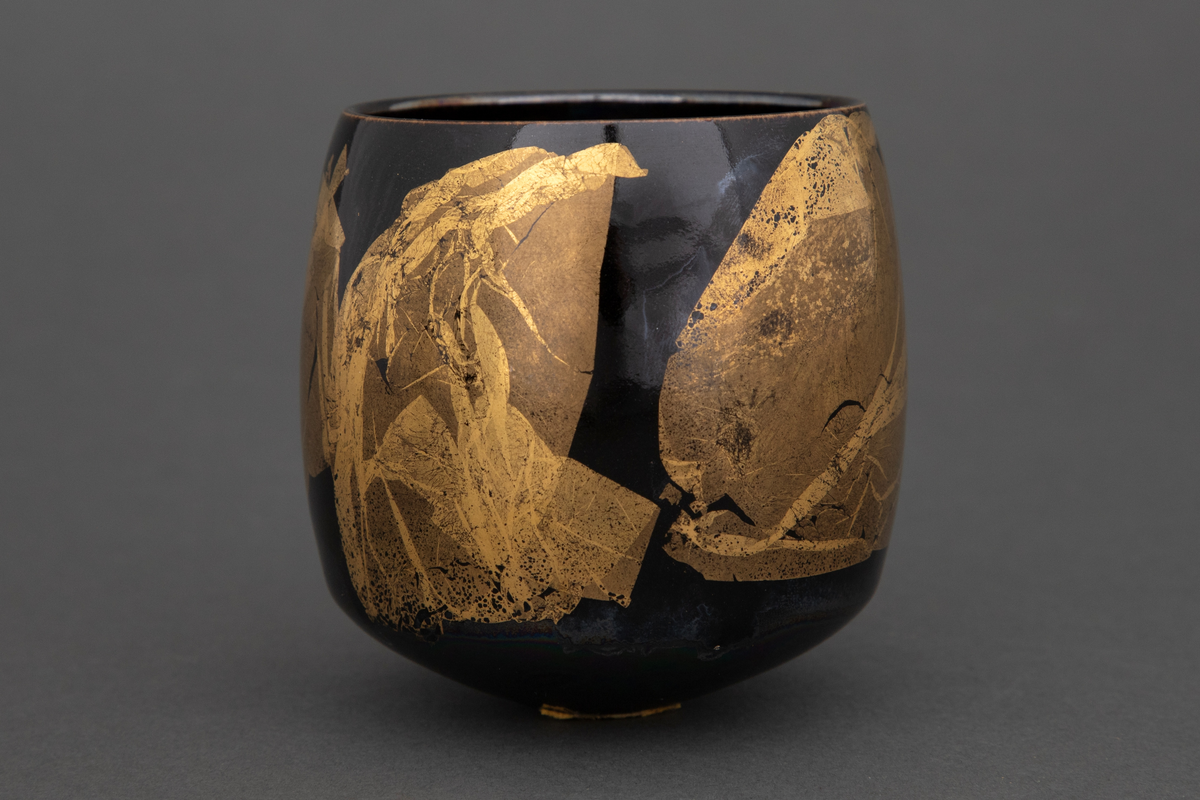 Bolle med liten bunnflate i porselen. Sylinderform som rundes av mot bunnen og toppen. Lilla, mørkeblå og grønn bunnkulør i bølgende formasjoner med uregelmessige former i bladgull utvendig.