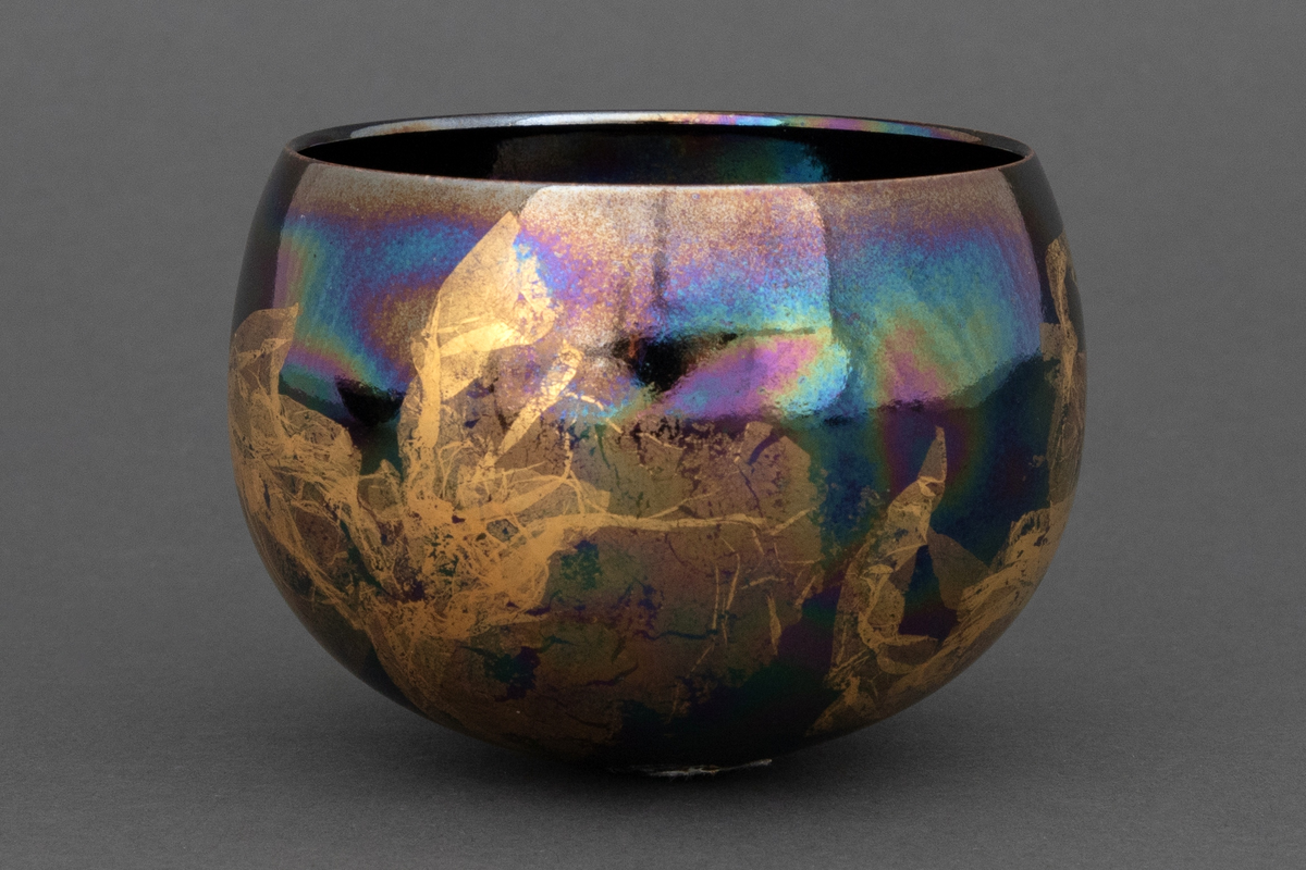 Bolle med liten bunnflate i porselen. Avrundet bunn og topp. Lilla, mørkeblå og grønn bunnkulør i bølgende formasjoner med uregelmessige former i bladgull utvendig.
