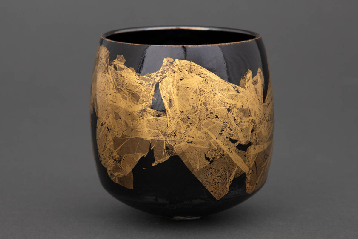 Bolle med liten bunnflate i porselen. Sylinderform som rundes av mot bunnen og toppen. Lilla, mørkeblå og grønn bunnkulør i bølgende formasjoner med uregelmessige former i bladgull utvendig.