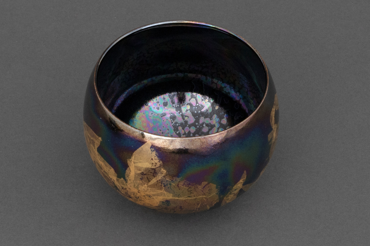 Bolle med liten bunnflate i porselen. Avrundet bunn og topp. Lilla, mørkeblå og grønn bunnkulør i bølgende formasjoner med uregelmessige former i bladgull utvendig.