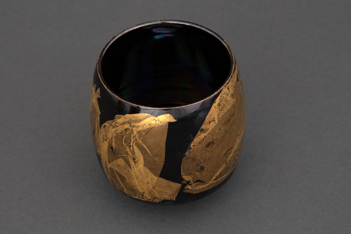 Bolle med liten bunnflate i porselen. Sylinderform som rundes av mot bunnen og toppen. Lilla, mørkeblå og grønn bunnkulør i bølgende formasjoner med uregelmessige former i bladgull utvendig.