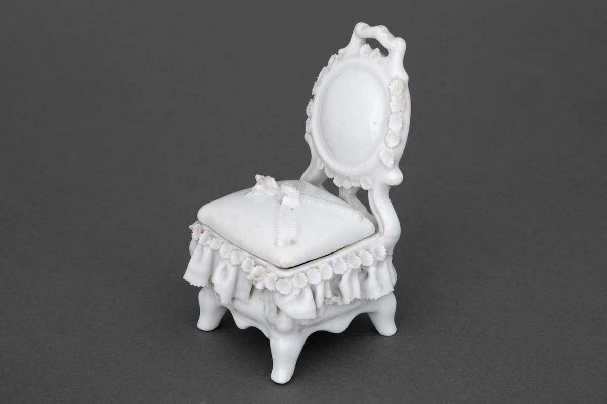 Stol i hvitglasert porselen med plass for oppbevaring under løs stolpute. Modellerte folder og små blomster rundt setet samt en blomsterbukett plassert på toppen av stolputen med spor etter forgylling.