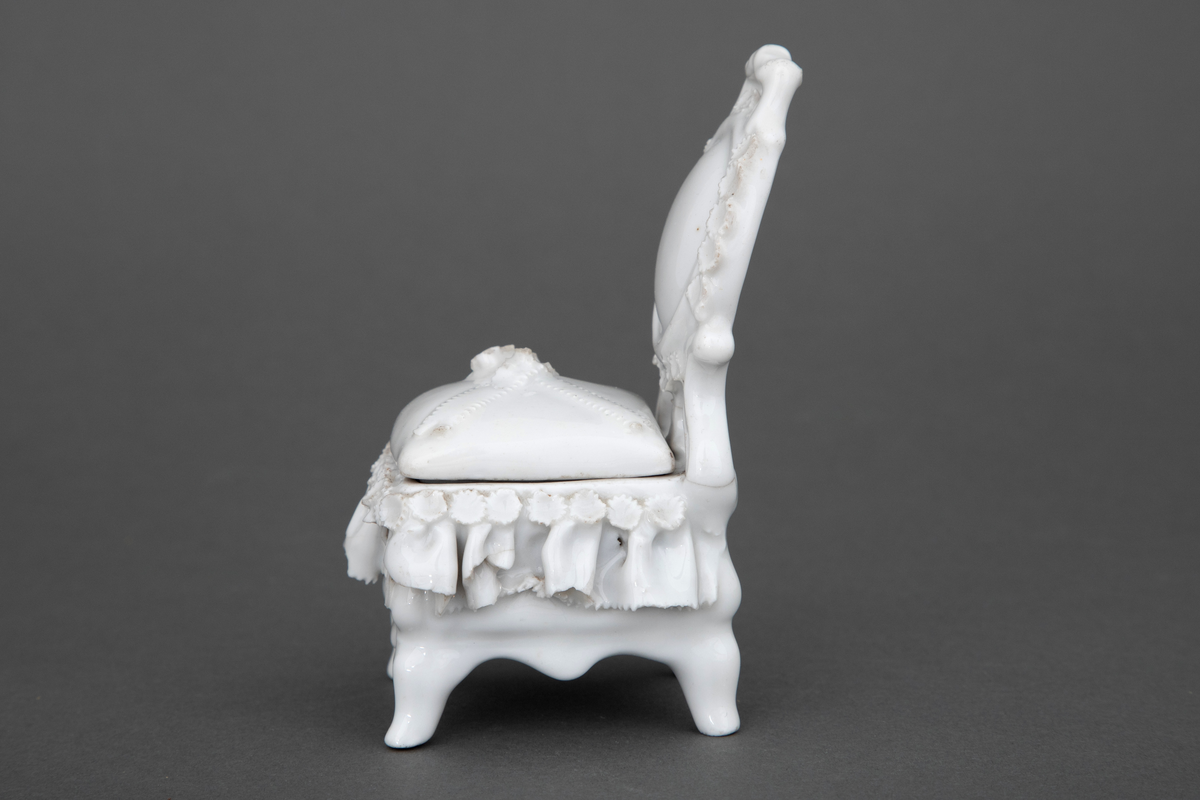 Stol i hvitglasert porselen med plass for oppbevaring under løs stolpute. Modellerte folder og små blomster rundt setet samt en blomsterbukett plassert på toppen av stolputen med spor etter forgylling.