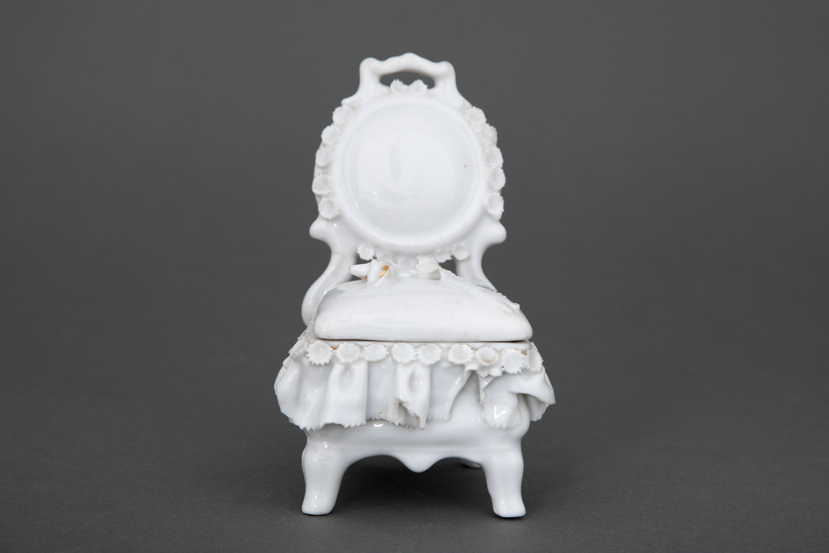Stol i hvitglasert porselen med plass for oppbevaring under løs stolpute. Modellerte folder og små blomster rundt setet samt en blomsterbukett plassert på toppen av stolputen med spor etter forgylling.