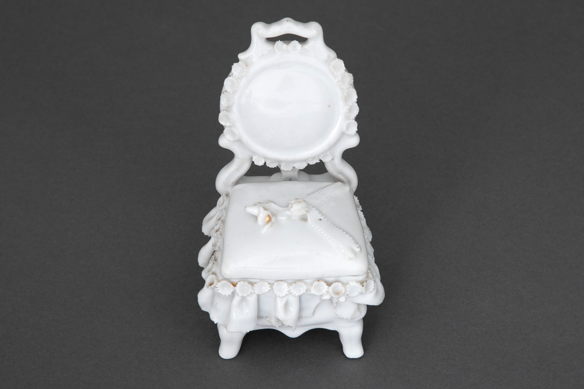 Stol i hvitglasert porselen med plass for oppbevaring under løs stolpute. Modellerte folder og små blomster rundt setet samt en blomsterbukett plassert på toppen av stolputen med spor etter forgylling.