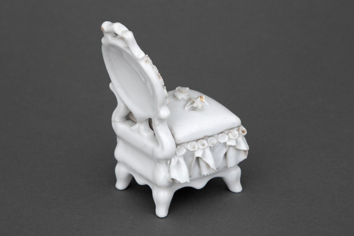 Stol i hvitglasert porselen med plass for oppbevaring under løs stolpute. Modellerte folder og små blomster rundt setet samt en blomsterbukett plassert på toppen av stolputen med spor etter forgylling.