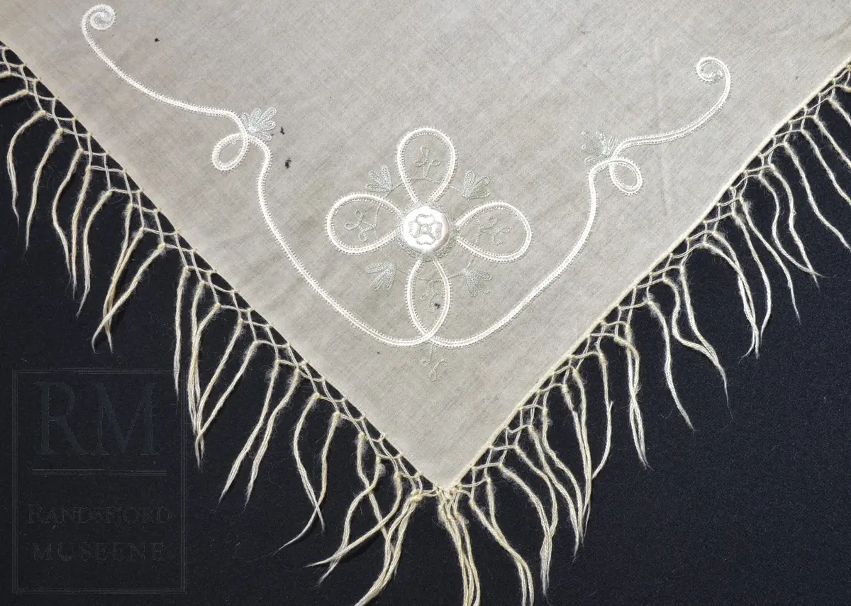 Kvadratisk, tynt ullstoff. Frynser langs alle fire kanter. Brodert motiv i et hjørne, antatt tamburering. 