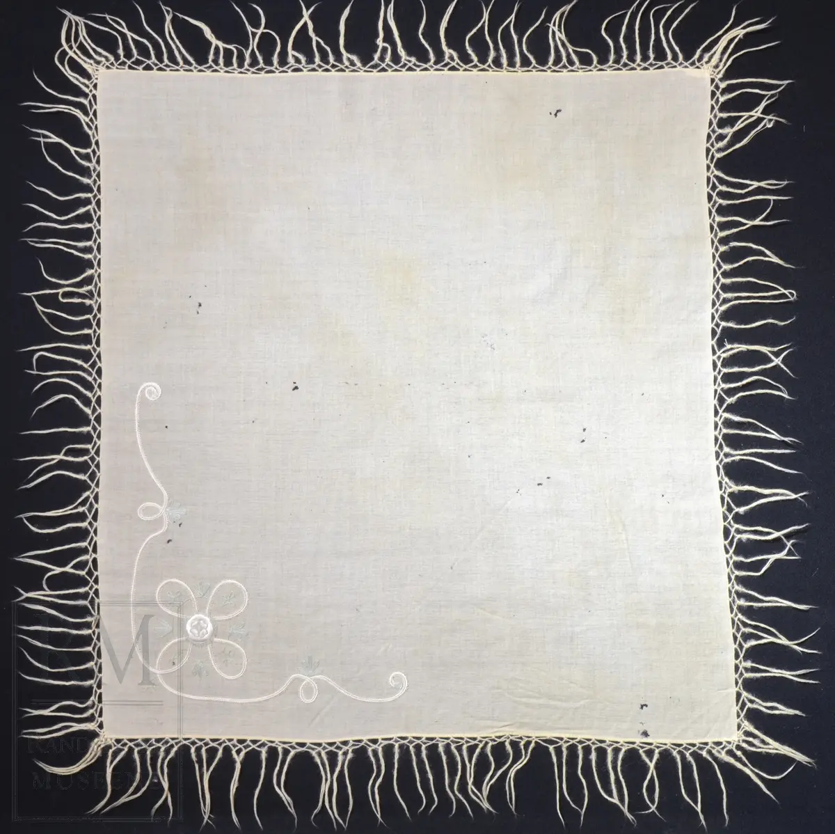 Kvadratisk, tynt ullstoff. Frynser langs alle fire kanter. Brodert motiv i et hjørne, antatt tamburering. 