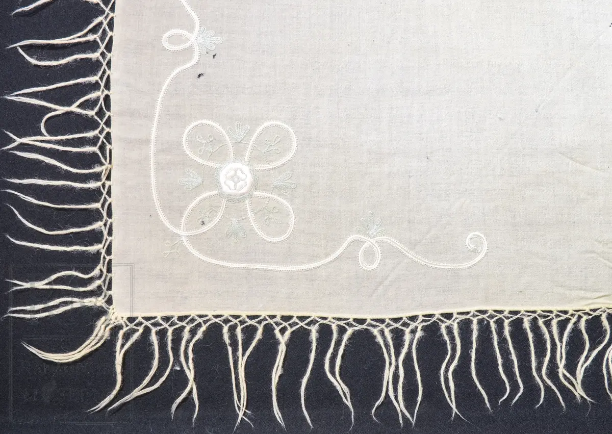 Kvadratisk, tynt ullstoff. Frynser langs alle fire kanter. Brodert motiv i et hjørne, antatt tamburering. 