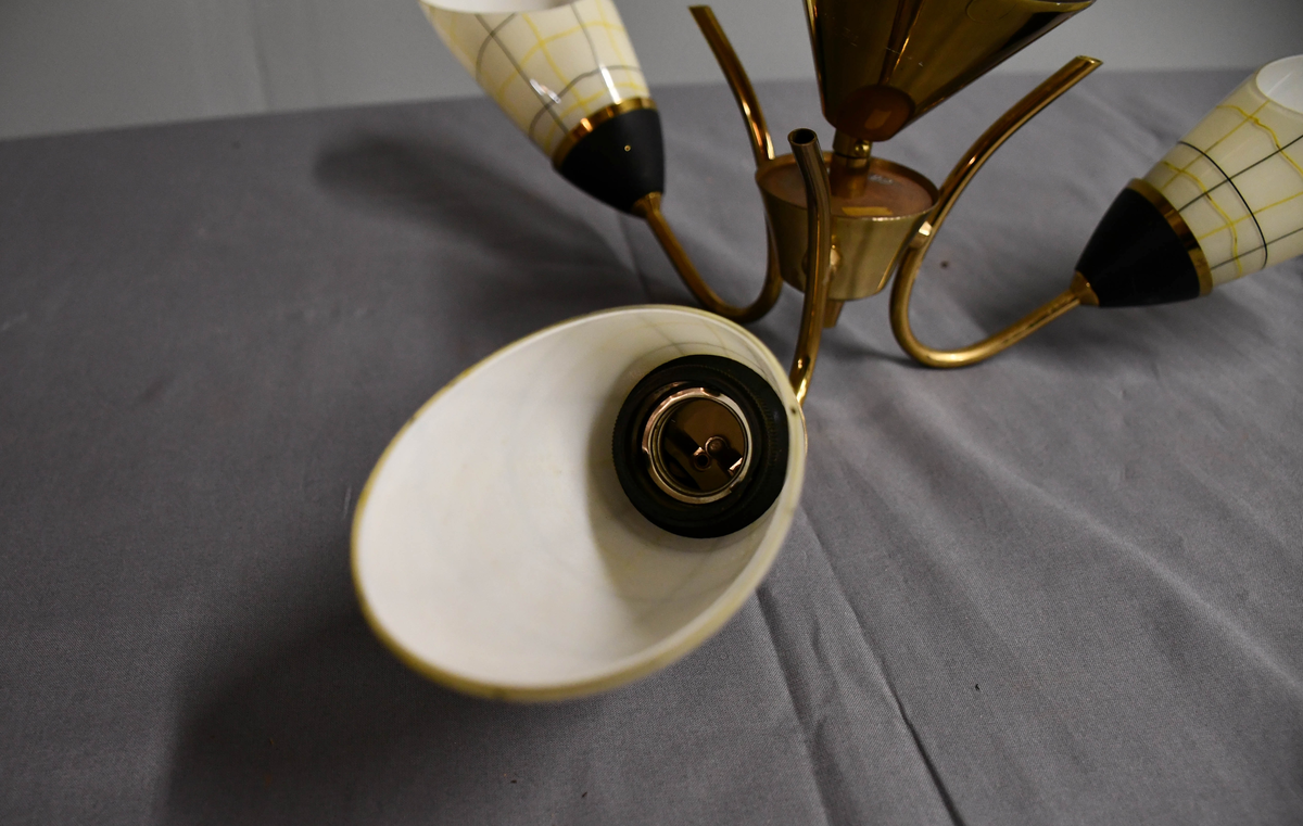 Trearmet hengelampe. Tynt messingrør med 'avkuttet kremmerhus' nede som feste for tre armer, og med påsatt messingskjuler for tilkobling oppe. Armene er krummet som tynne stilker og holder hver en elektrisk fatning med en 'snøklokke'- skjerm: en skrått avkuttet glassdråpe med et sort/gult nett streket opp med 'usikker' hånd, og med sort-eloksert messingbeger . Skjermene er invendig hvite og utvendig lyst benhvite. 'Blomstene' 'henger ned-opp' så de kaster lyset opp i taket.