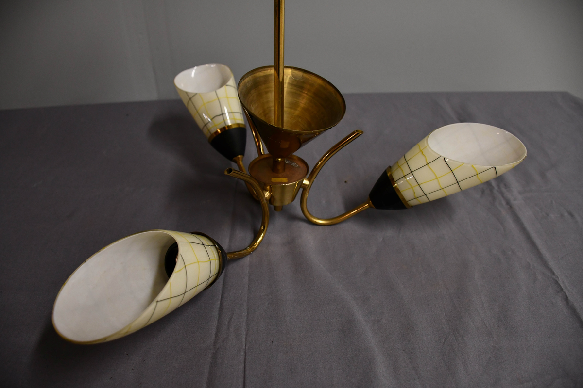 Trearmet hengelampe. Tynt messingrør med 'avkuttet kremmerhus' nede som feste for tre armer, og med påsatt messingskjuler for tilkobling oppe. Armene er krummet som tynne stilker og holder hver en elektrisk fatning med en 'snøklokke'- skjerm: en skrått avkuttet glassdråpe med et sort/gult nett streket opp med 'usikker' hånd, og med sort-eloksert messingbeger . Skjermene er invendig hvite og utvendig lyst benhvite. 'Blomstene' 'henger ned-opp' så de kaster lyset opp i taket.