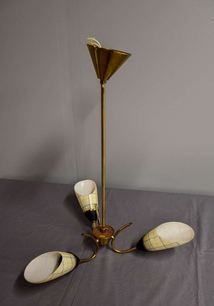 Trearmet hengelampe. Tynt messingrør med 'avkuttet kremmerhus' nede som feste for tre armer, og med påsatt messingskjuler for tilkobling oppe. Armene er krummet som tynne stilker og holder hver en elektrisk fatning med en 'snøklokke'- skjerm: en skrått avkuttet glassdråpe med et sort/gult nett streket opp med 'usikker' hånd, og med sort-eloksert messingbeger . Skjermene er invendig hvite og utvendig lyst benhvite. 'Blomstene' 'henger ned-opp' så de kaster lyset opp i taket.