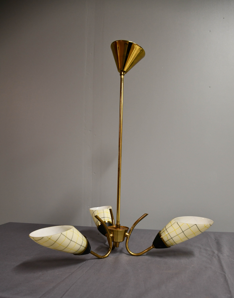 Trearmet hengelampe. Tynt messingrør med 'avkuttet kremmerhus' nede som feste for tre armer, og med påsatt messingskjuler for tilkobling oppe. Armene er krummet som tynne stilker og holder hver en elektrisk fatning med en 'snøklokke'- skjerm: en skrått avkuttet glassdråpe med et sort/gult nett streket opp med 'usikker' hånd, og med sort-eloksert messingbeger . Skjermene er invendig hvite og utvendig lyst benhvite. 'Blomstene' 'henger ned-opp' så de kaster lyset opp i taket.