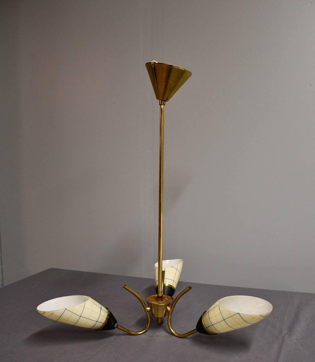 Trearmet hengelampe. Tynt messingrør med 'avkuttet kremmerhus' nede som feste for tre armer, og med påsatt messingskjuler for tilkobling oppe. Armene er krummet som tynne stilker og holder hver en elektrisk fatning med en 'snøklokke'- skjerm: en skrått avkuttet glassdråpe med et sort/gult nett streket opp med 'usikker' hånd, og med sort-eloksert messingbeger . Skjermene er invendig hvite og utvendig lyst benhvite. 'Blomstene' 'henger ned-opp' så de kaster lyset opp i taket.