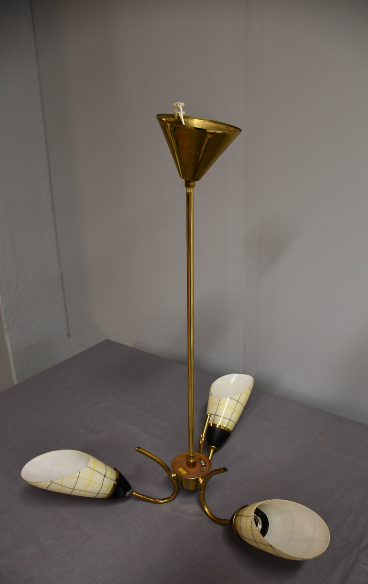 Trearmet hengelampe. Tynt messingrør med 'avkuttet kremmerhus' nede som feste for tre armer, og med påsatt messingskjuler for tilkobling oppe. Armene er krummet som tynne stilker og holder hver en elektrisk fatning med en 'snøklokke'- skjerm: en skrått avkuttet glassdråpe med et sort/gult nett streket opp med 'usikker' hånd, og med sort-eloksert messingbeger . Skjermene er invendig hvite og utvendig lyst benhvite. 'Blomstene' 'henger ned-opp' så de kaster lyset opp i taket.