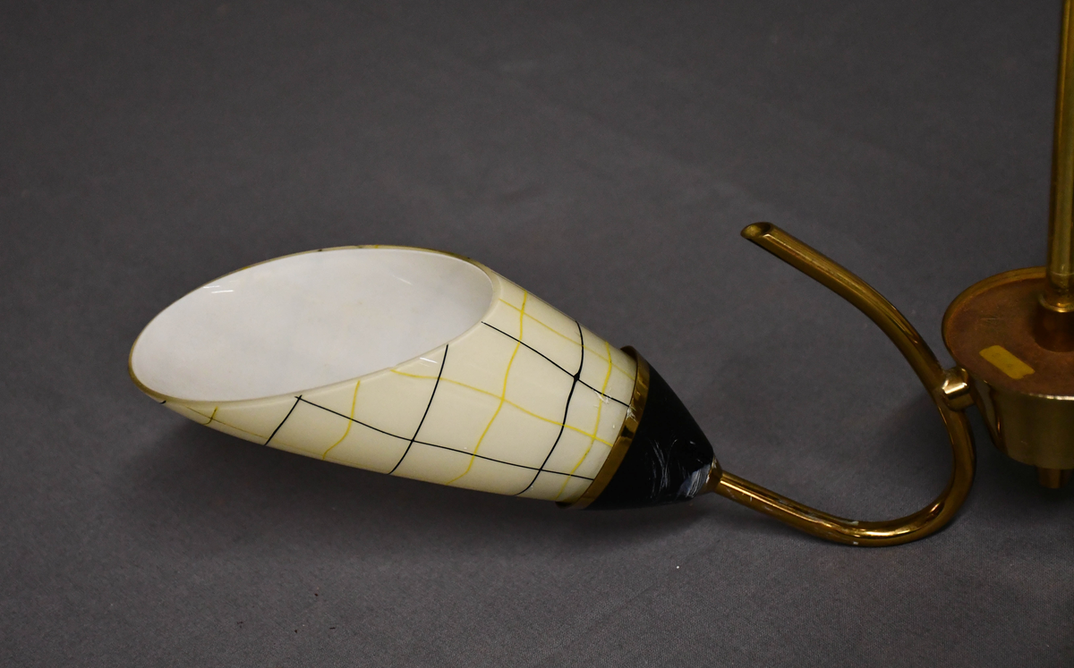 Trearmet hengelampe. Tynt messingrør med 'avkuttet kremmerhus' nede som feste for tre armer, og med påsatt messingskjuler for tilkobling oppe. Armene er krummet som tynne stilker og holder hver en elektrisk fatning med en 'snøklokke'- skjerm: en skrått avkuttet glassdråpe med et sort/gult nett streket opp med 'usikker' hånd, og med sort-eloksert messingbeger . Skjermene er invendig hvite og utvendig lyst benhvite. 'Blomstene' 'henger ned-opp' så de kaster lyset opp i taket.