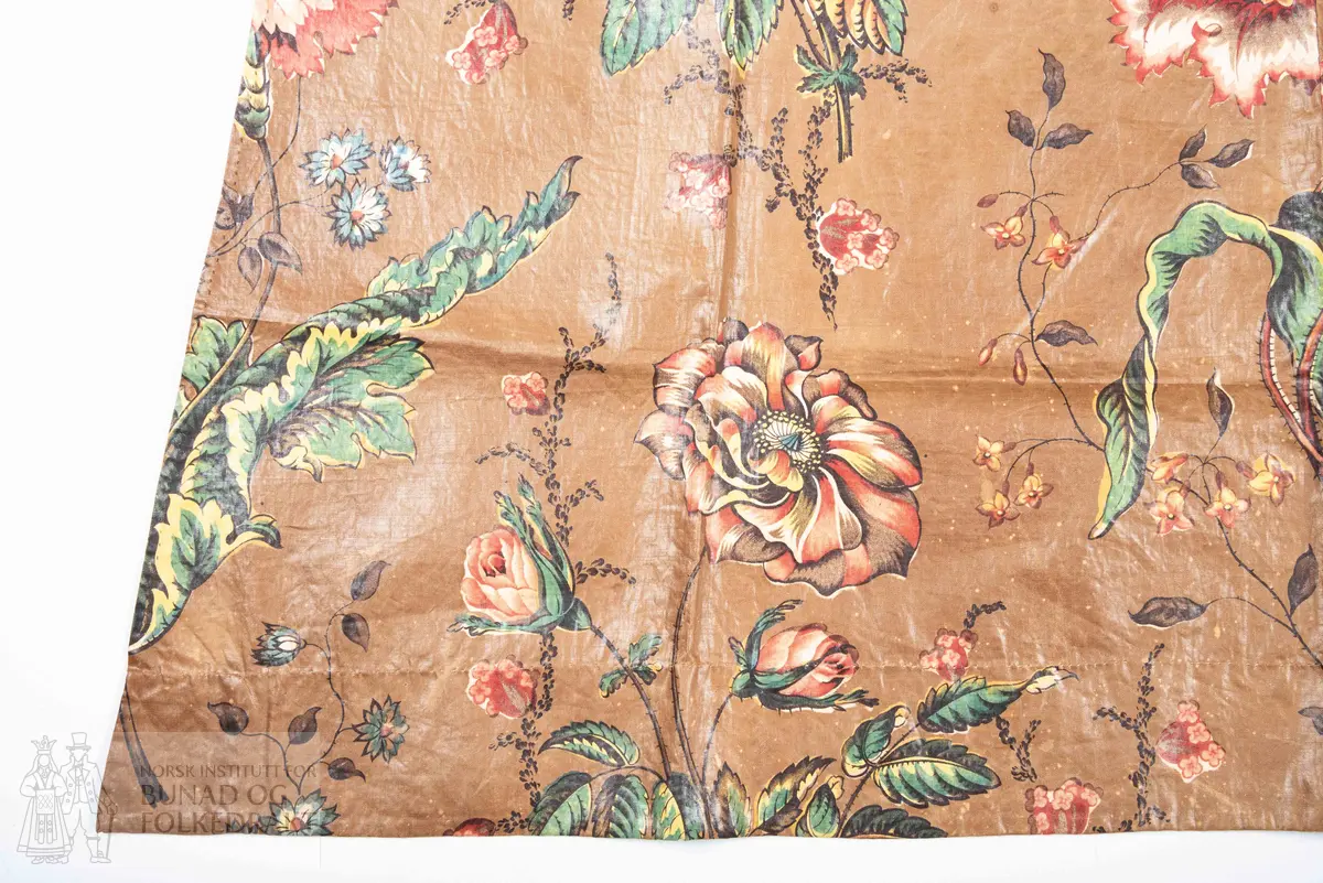 Forkle.
Forkledet er omsydd.
Trykt, glansfullt chintz. Saum midt foran, attersting og overkasting med lyst silkegarn.
Skonert langs nedkanten med eit 5 cm breitt, trykt bomullstøy.
Dette er falda til langs skoneringas øvre kant. Nederst er det bretta dobbel, smal fald på forkletøyet, som er falda til i skoneringa.
Sidene er truleg sekundære då dei er skråklypte. Dei har dobbel smal fald, sauma til med små faldesting, men trådtypa er annleis og yngre.
I livet er det bretta ned ca 1 cm som er stikka ned med ein maskinsaum.