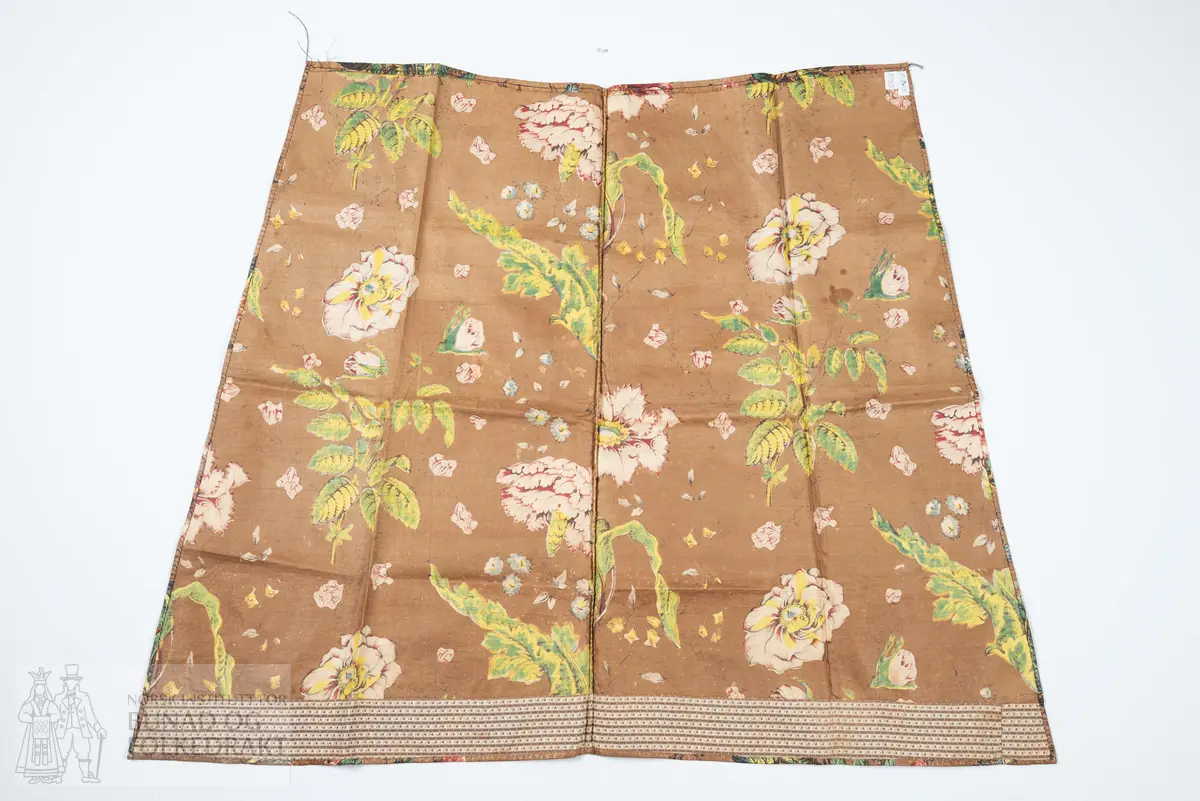 Forkle.
Forkledet er omsydd.
Trykt, glansfullt chintz. Saum midt foran, attersting og overkasting med lyst silkegarn.
Skonert langs nedkanten med eit 5 cm breitt, trykt bomullstøy.
Dette er falda til langs skoneringas øvre kant. Nederst er det bretta dobbel, smal fald på forkletøyet, som er falda til i skoneringa.
Sidene er truleg sekundære då dei er skråklypte. Dei har dobbel smal fald, sauma til med små faldesting, men trådtypa er annleis og yngre.
I livet er det bretta ned ca 1 cm som er stikka ned med ein maskinsaum.