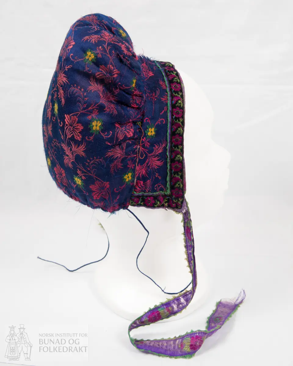 Rynkelue, kvadratisk pull rynka til eit rektangulært pannestykkje. Hovudstoff av satengvove blomemønstra silkestoff. Fòr av stripevove bomullslerret. Pullstykkjet er kraftig vattert med mjuk tjukk vatt, medan pannestykkjet er utan mellomfor. Løpegang med trekksnor i nedkant av pullstykkjet. Knyteband av bomull/silke, med vove blomemønster. Dette bandet er også brukt til kanting av pannestykkjet. Oppå dette er sydd blomemønstra fløyelsband. Heile lua er handsydd.