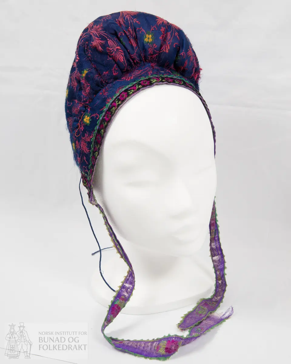 Rynkelue, kvadratisk pull rynka til eit rektangulært pannestykkje. Hovudstoff av satengvove blomemønstra silkestoff. Fòr av stripevove bomullslerret. Pullstykkjet er kraftig vattert med mjuk tjukk vatt, medan pannestykkjet er utan mellomfor. Løpegang med trekksnor i nedkant av pullstykkjet. Knyteband av bomull/silke, med vove blomemønster. Dette bandet er også brukt til kanting av pannestykkjet. Oppå dette er sydd blomemønstra fløyelsband. Heile lua er handsydd.