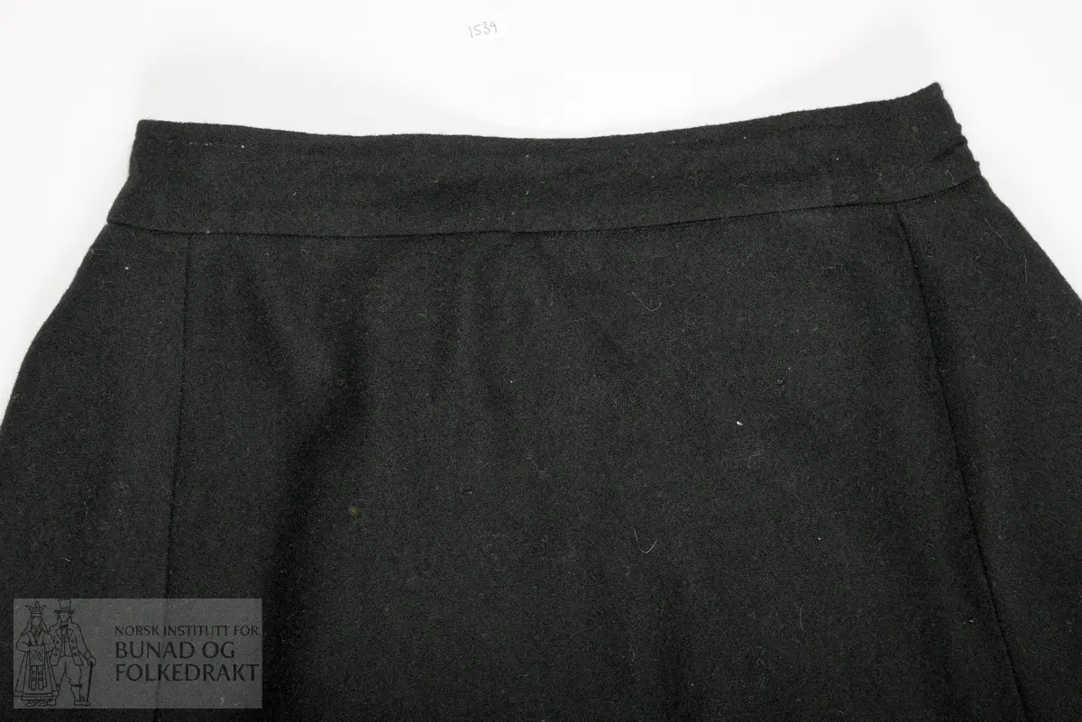 Stakk sauma av godt valka svart vadmål. Samansett av tre lett utskrådde høgder framme, og to rette høgder bak. Splitt oppe mb, og lukking med hekter i linning. På kvar side av splitt mb er stakken lagt i eit felt med 6 smale svært djupe faldar, som alle vender mot mb. I det faldelagte feltet er stakkelengda 2 cm kortare enn rundt resten av stakken. Elles er stakken sydd glatt til linning. Linningen er sydd med maskinsaum. Høgdene framme er sydd med maskinsaum, og høgdene bak er sydd med handsaum med lintråd, sydd med lange attersting som syner som prikk på eine sida. Skoning og kanting nede er sydd for hand med gråbrun lintråd. På retta etter nedkanten er påsydd eit stoff av brun bomullsfløyel. Og ovanfor denne er påsydd ein smal blå kroklisse av ull. På innsida er sydd på ei skoning av svart godt valka vadmål. Denne er sydd til rette mot rette, så lagt rundt på innsida, med ompaspolering, slik at vadmålet blir som ein tittekant nedst.