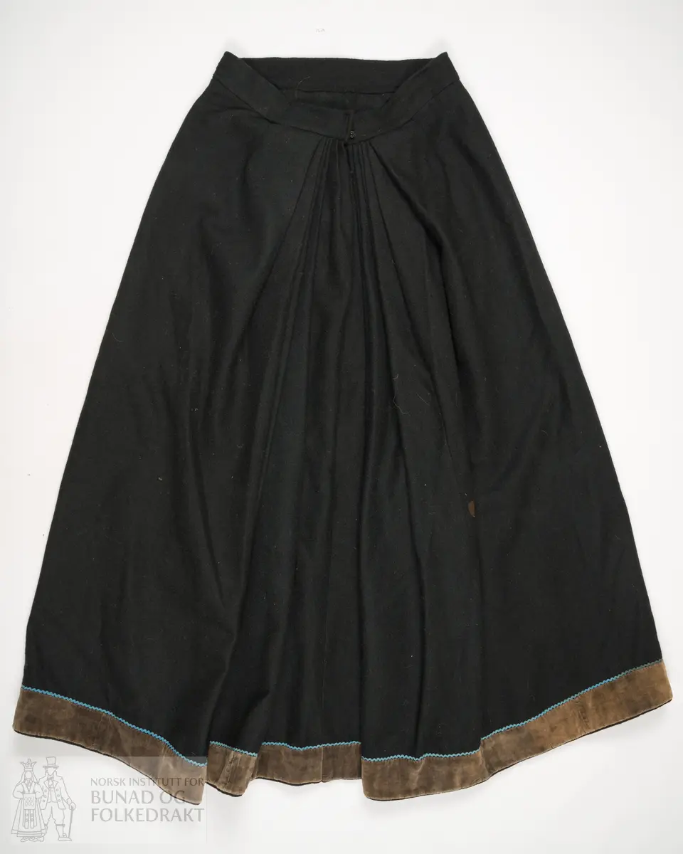 Stakk sauma av godt valka svart vadmål. Samansett av tre lett utskrådde høgder framme, og to rette høgder bak. Splitt oppe mb, og lukking med hekter i linning. På kvar side av splitt mb er stakken lagt i eit felt med 6 smale svært djupe faldar, som alle vender mot mb. I det faldelagte feltet er stakkelengda 2 cm kortare enn rundt resten av stakken. Elles er stakken sydd glatt til linning. Linningen er sydd med maskinsaum. Høgdene framme er sydd med maskinsaum, og høgdene bak er sydd med handsaum med lintråd, sydd med lange attersting som syner som prikk på eine sida. Skoning og kanting nede er sydd for hand med gråbrun lintråd. På retta etter nedkanten er påsydd eit stoff av brun bomullsfløyel. Og ovanfor denne er påsydd ein smal blå kroklisse av ull. På innsida er sydd på ei skoning av svart godt valka vadmål. Denne er sydd til rette mot rette, så lagt rundt på innsida, med ompaspolering, slik at vadmålet blir som ein tittekant nedst.