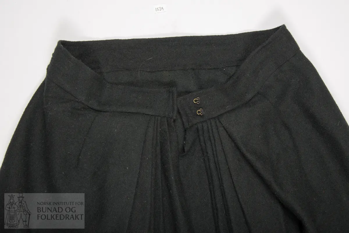 Stakk sauma av godt valka svart vadmål. Samansett av tre lett utskrådde høgder framme, og to rette høgder bak. Splitt oppe mb, og lukking med hekter i linning. På kvar side av splitt mb er stakken lagt i eit felt med 6 smale svært djupe faldar, som alle vender mot mb. I det faldelagte feltet er stakkelengda 2 cm kortare enn rundt resten av stakken. Elles er stakken sydd glatt til linning. Linningen er sydd med maskinsaum. Høgdene framme er sydd med maskinsaum, og høgdene bak er sydd med handsaum med lintråd, sydd med lange attersting som syner som prikk på eine sida. Skoning og kanting nede er sydd for hand med gråbrun lintråd. På retta etter nedkanten er påsydd eit stoff av brun bomullsfløyel. Og ovanfor denne er påsydd ein smal blå kroklisse av ull. På innsida er sydd på ei skoning av svart godt valka vadmål. Denne er sydd til rette mot rette, så lagt rundt på innsida, med ompaspolering, slik at vadmålet blir som ein tittekant nedst.