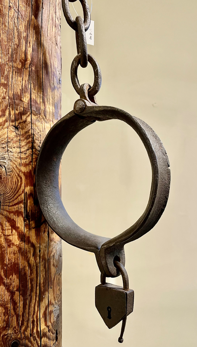 Brune trestokk med enkel utskåret profilering i form av runde bånd øverst og nedert. Påmontert halsjern med lenke og hengelås.