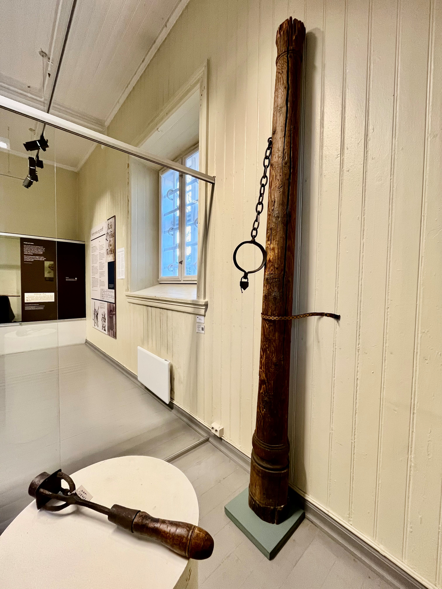 Brune trestokk med enkel utskåret profilering i form av runde bånd øverst og nedert. Påmontert halsjern med lenke og hengelås.