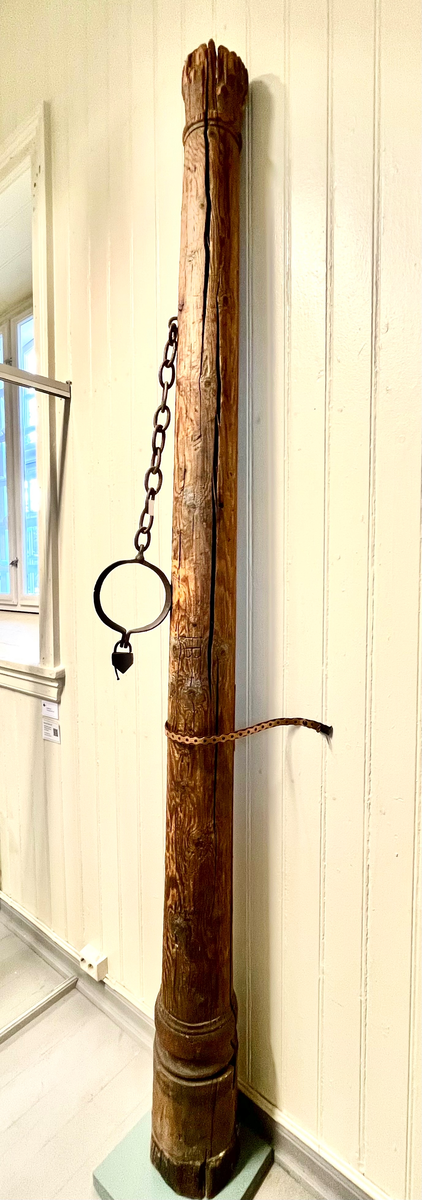 Brune trestokk med enkel utskåret profilering i form av runde bånd øverst og nedert. Påmontert halsjern med lenke og hengelås.