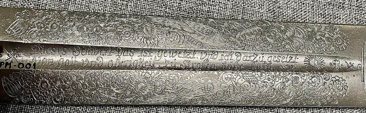 Langt tveegget sverd i stål med rett, butt klinge med kort blodrand på hver side. Klingen er rikt utsmykket med etsinger eller inngraveringer og flere merker som kan være smedmerker, blant annet en sol med menneskeansikt. Dekorert med blomsterranker, fugle- og dyremotiver og gudinnen Justitia. Grepet har hjerteformet knapp avsluttet med en mindre knapp. Stålgrep viklet med tvunnet messingtråd og satt med messingbånd oppe og nede. Symetrisk feste med flate pilformede parerstenger som smalner inn mot midten.