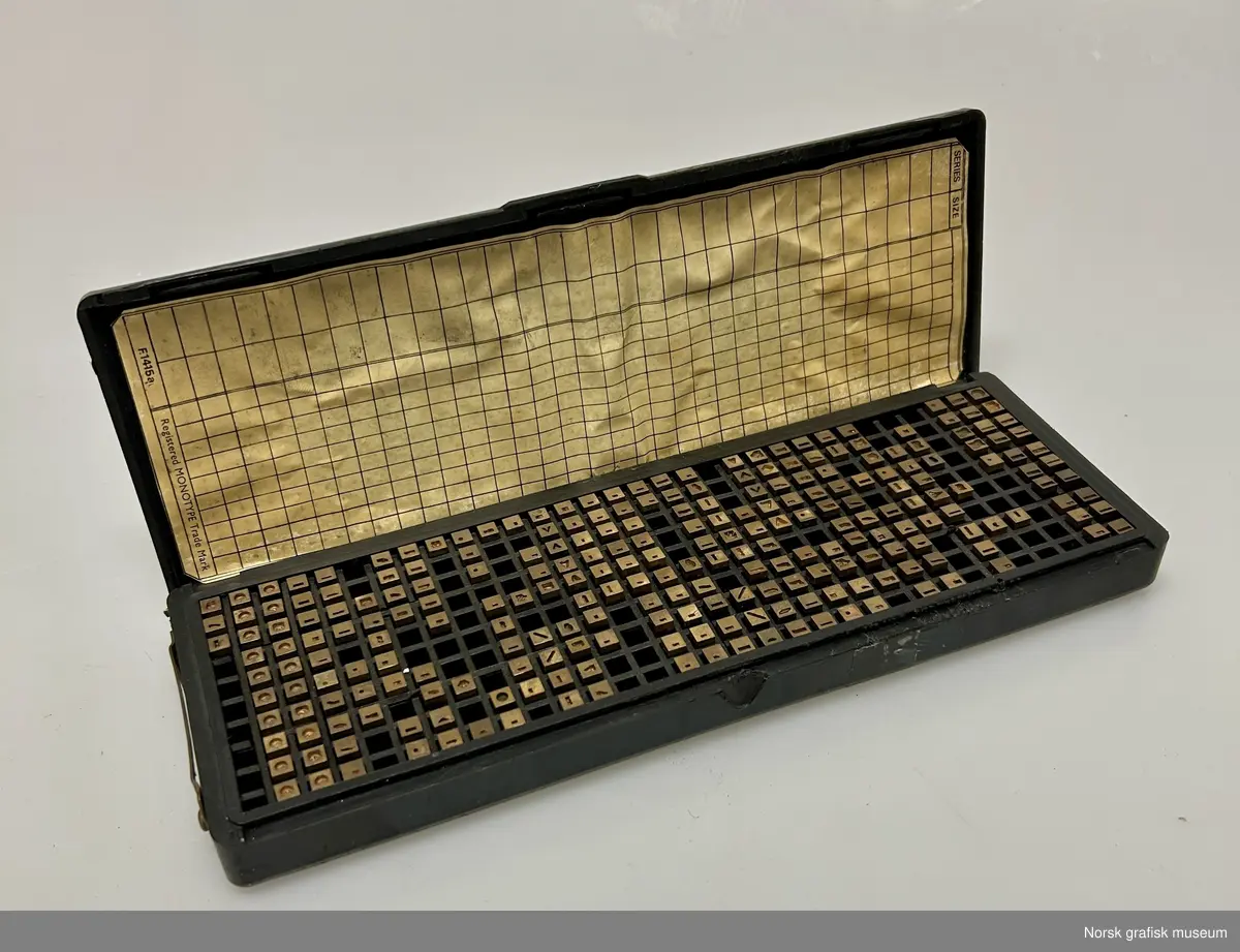 Et plastskrin inneholdende matriser til Monotype. De individuelle matrisene er plassert i små rom i skrinet. På undersiden av lokket et det limt et skjema. Firmanavet er preget inn i lokket.